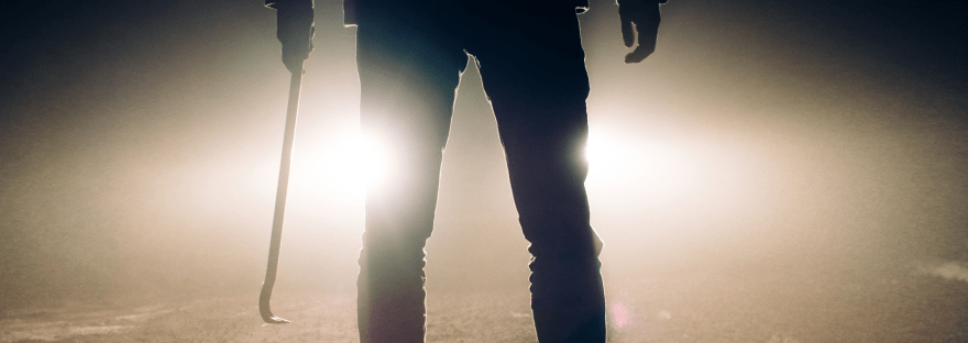 Photograph of a man silhouetted by car headlights, holding a crowbar.